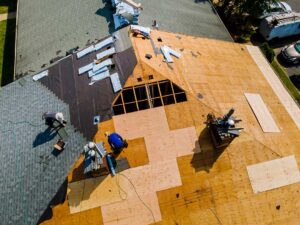 A roofing crew removes old shingles from a residential roof using pitchforks and places debris into a nearby dumpster.